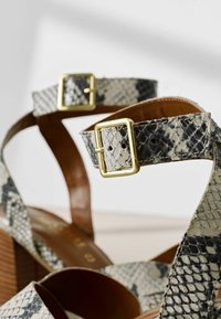 Sandali con tacco in motivo pelle di serpente grigio e beige. Presentano una fibbia dorata e dettagli in pelle marrone. Tacco in legno.