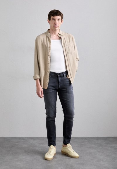 Camisa de botón beige sobre una camiseta sin mangas blanca, combinada con jeans oscuros y zapatillas amarillas claras con un acabado texturizado. Diseño simple en general.