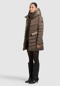 Hnedý prešívaný kabát s vysokým golierom, vertikálnym prešívaným vzorom, bočnými vreckami a čiernou logovou známkou. Nosený s vysokými čiernymi koženými čižmami.