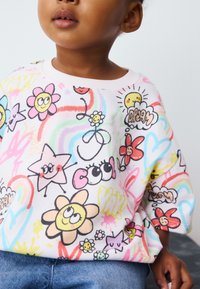 Sweatshirt en coton blanc avec des motifs cartoon colorés et ludiques de fleurs, d'étoiles et de visages souriants, doté d'un col rond et de manches courtes.