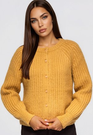 Une femme pose devant un fond gris clair, vêtue d’un cardigan en maille jaune moutarde boutonné, les mains jointes à la taille.