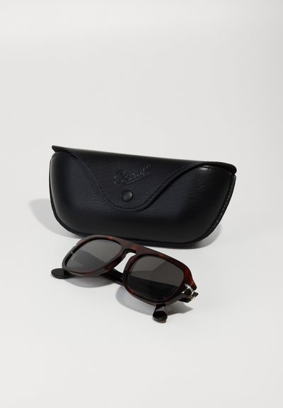 Óculos de sol com lentes escuras numa armação de tartaruga, ao lado de uma caixa de couro preta em relevo com a marca "Persol".