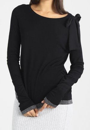 Haut noir à manches longues avec un col en rond, présentant un détail en noeud sur l'épaule gauche et un ourlet accentué de silver. Tissu doux et extensible.