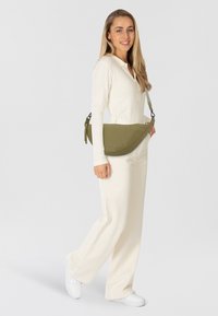 Donna in abbigliamento crema e sneaker bianche sorride, portando una borsa a tracolla verde oliva su uno sfondo bianco semplice.