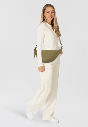 Donna in abbigliamento crema e sneaker bianche sorride, portando una borsa a tracolla verde oliva su uno sfondo bianco semplice.