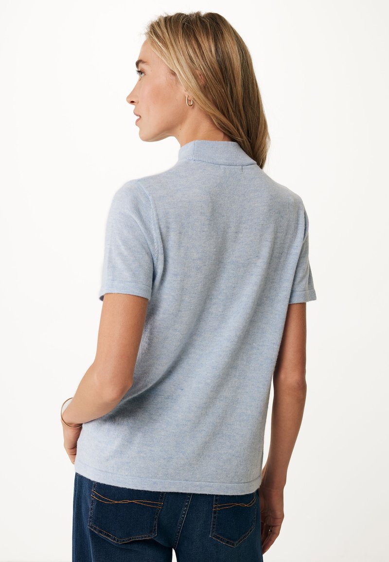 Kobieta z długimi blond włosami, ubrana w jasnoniebieski sweter z krótkim rękawem i ciemnoniebieskie dżinsy, stojąca w profilu na białym tle.