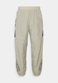 Světle béžové teplákové kalhoty s elastickým pasem, černými bočními panely a bílým brandingem na dolních nohavicích. Vyrobeno z lehkého materiálu.