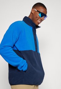 Blauer und marineblauer Fleece-Pullover mit hohem Kragen, knöpfbarem Frontverschluss und seitlichen Taschen, der eine weiche Textur und ein kontrastierendes Farbmuster aufweist.