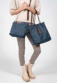 Two blue fabric tote bags with brown leather straps, featuring a minimalist design and a prominent tassel detail. Worn with checkered pants.