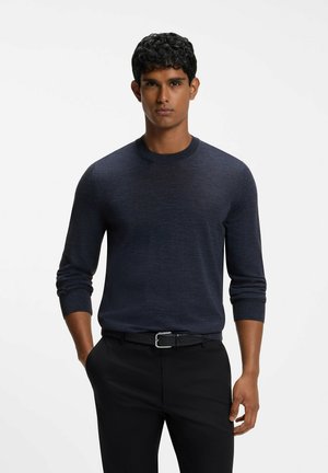 Mand med krøllet mørkt hår iført en marineblå langærmet sweater stoppet ned i sorte bukser med sort bælte, stående med den ene hånd i lommen.