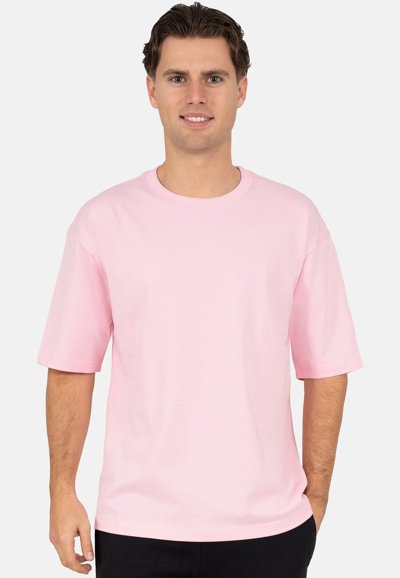 T-shirt à manches courtes et col rond en tissu uni rose. Présente une coupe décontractée et une texture lisse, portée avec un pantalon foncé.