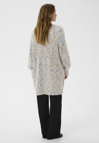 Frau mit langen braunen Haaren, die einen langen, gepunkteten Strickcardigan und schwarze, weit geschnittene Hosen trägt, steht mit dem Rücken zu einer weißen Wand.