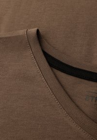 Camiseta marrón con un clásico cuello redondo, que presenta una textura de tela suave y costuras negras en contraste a lo largo del cuello.