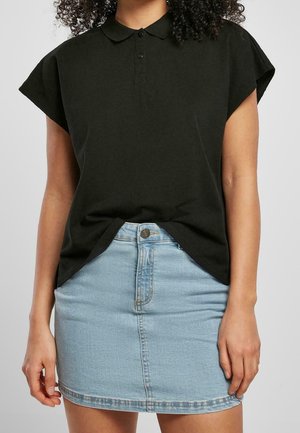Vrouw die een zwart poloshirt met korte mouwen draagt, lichtjes ingestopt in een lichtblauwe denim minirok, staand tegen een effen achtergrond.