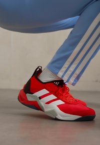 Baskets de sport rouges avec des rayures blanches, en tissu texturé, avec un col rembourré et une semelle épaisse. Amorcé avec un pantalon de survêtement bleu clair.