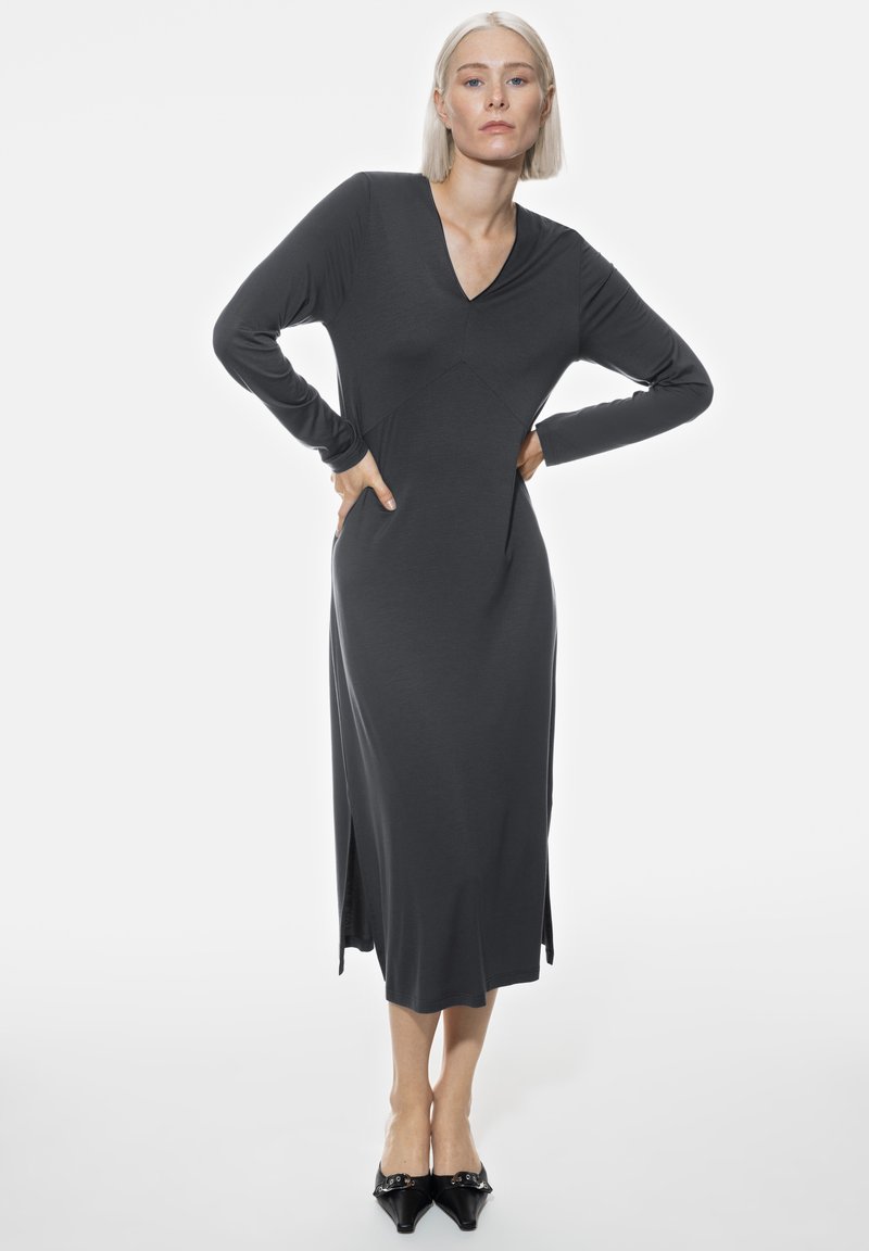 Lange, getailleerde jurk van antracietgrijze stof. Heeft een V-hals, lange mouwen, zijsplitsen en een slanke pasvorm; gecombineerd met zwarte hakken.