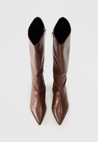 Une paire de bottes en cuir marron hautes avec des bouts pointus et des fermetures éclair sur le côté, posées verticalement sur une surface blanche.