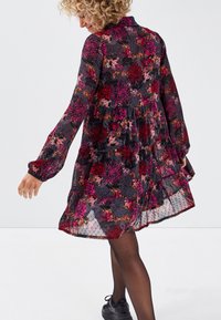 Robe florale à manches longues, motif noir et bordeaux sur tissu transparent, design à volants et ourlet à volants. Portée avec des baskets noires.