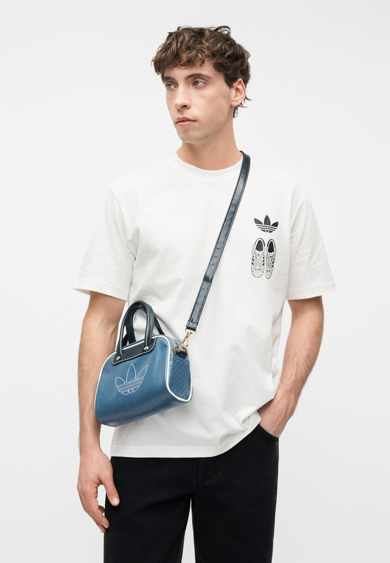Giovane uomo con t-shirt bianca Adidas con logo e stampa di sneaker, indossa una borsa a tracolla blu Adidas e pantaloni neri, in piedi contro uno sfondo bianco.