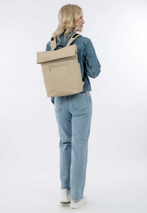 Sac à dos beige à rabat avec des bretelles réglables, texture lisse et poche avant. Porté avec une veste en jean et un jean bleu clair.