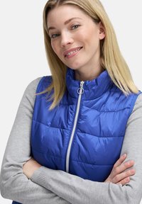 Blauer Steppweste mit hohem Kragen und durchgehendem Reißverschluss, ausgestattet mit einem gesteppten Design. Über einem grauen Langarmshirt getragen. Glattes, glänzendes Material.