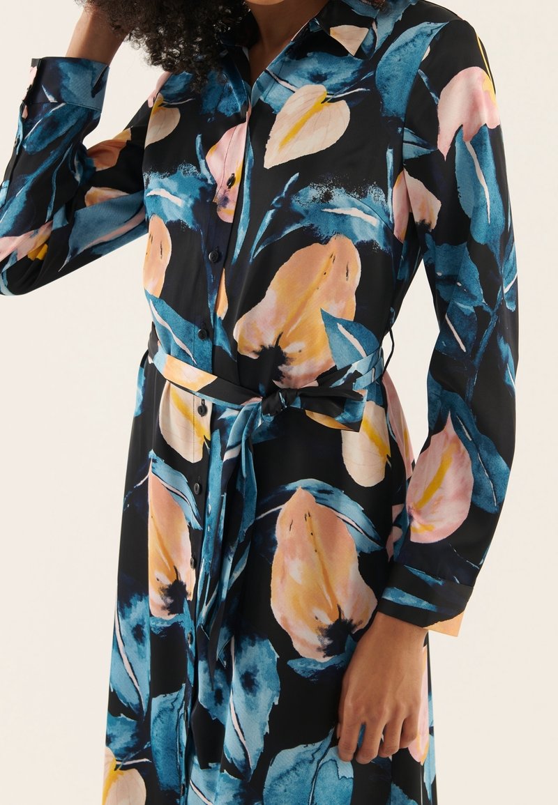 Robe chemise fleurie à manches longues, tissu noir orné de grandes fleurs roses et bleues, boutons sur le devant, et ceinture nouée à la taille.