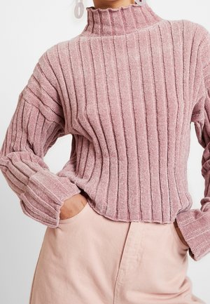 Femme portant un pull côtelé rose poussiéreux à manches longues avec un col haut et un pantalon rose clair, mains dans les poches.