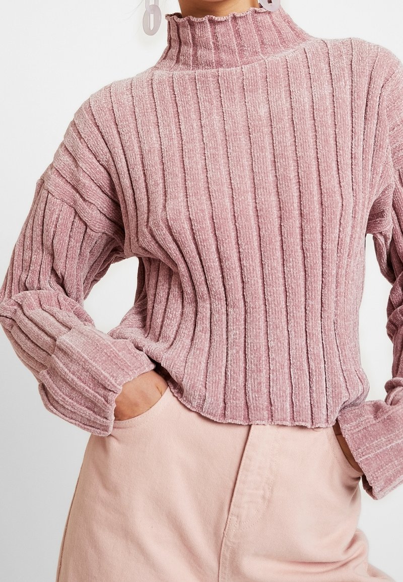 Femme portant un pull côtelé rose poussiéreux à manches longues avec un col haut et un pantalon rose clair, mains dans les poches.