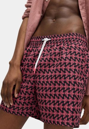 Primo piano di una persona che indossa pantaloncini rosa con motivo a fantasia e coulisse bianca, e una camicia rosa a maniche lunghe arrotolata, che mostra l'addome tonico e la mano.