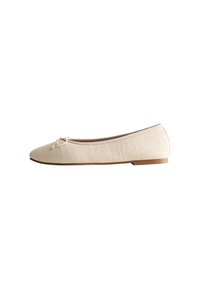 Zapato plano beige con parte superior de tela texturizada, puntera redondeada y un lazo decorativo. Presenta una suela de goma marrón baja para mayor comodidad.