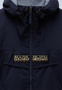 Granatowa kurtka z teksturowaną szarą podszewką, wyposażona w przedni zamek błyskawiczny oraz dwa wyraźne naszywki z logo w kontrastujących kolorach.