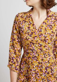 Robe à fleurs en jaune moutarde avec un design en portefeuille. Présente des fleurs violettes, roses et vertes, un col et des manches trois-quarts.