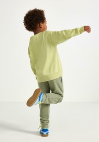 Lys grøn sweatshirt og olivengrønne joggingbukser, parret med blå og grønne sneakers. Barnet står på ét ben med en arm hævet.