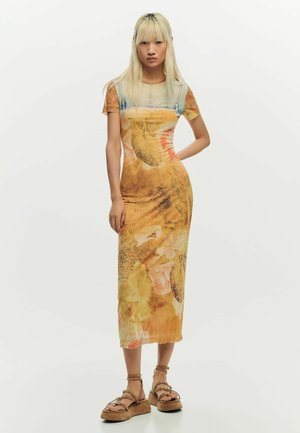 Noor naine pikkade blondide juustega kannab keha järgset, oranži ja beeži mustriga midi-kleiti ning pruune platvormsandaale, seistes valge tausta ees.