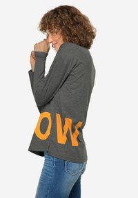 Maglietta grigia a maniche lunghe con grande grafica arancione "OW" sul lato, tessuto leggero, vestibilità comoda e spacchi laterali per una maggiore libertà di movimento.