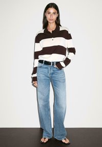 Striped long-sleeve polo shirt in brown and cream, paired with light blue wide-leg jeans and black belt. Model stands on a neutral background.
