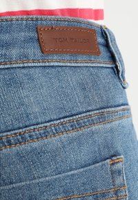 Jeans de mezclilla azules con parche de cuero marrón etiquetado "TOM TAILOR", costuras visibles y diseño tradicional de bolsillo trasero.