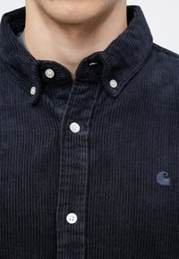 Ναυτική καμπαρντίνα με γιακά που κλείνει με κουμπιά, διαθέτει λευκά κουμπιά και ένα μικρό κεντημένο λογότυπο στο αριστερό στήθος. Υφασμάτινη βελούδινη υφή.