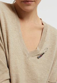 Beige sweater with a deep V-neck and ribbed edges featuring a decorative safety pin accent with black and pink beads.