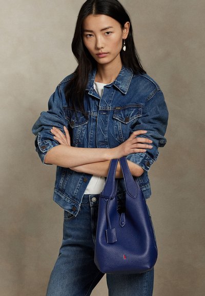 Kvinde med langt mørkt hår iført en denimjakke og jeans, der holder en textured blå håndtaske med krydsede arme mod en ensfarvet baggrund.