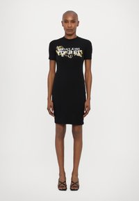 Juoda prigludusi suknelė su trumpomis rankovėmis, su dekoratyviniu baltu ir auksiniu "Versace Jeans Couture" logotipu ant krūtinės.