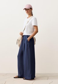 Femeie purtând o șapcă roz, bluză albă cu mâneci scurte, pantaloni largi bleumarin, sandale negre și având o geantă bej purtată în diagonală, stând pe un podea cu gresie.