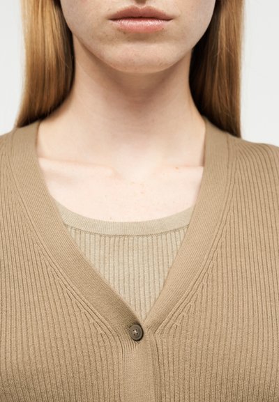 Κοντινό πλάνο στον λαιμό και τα χείλη ενός ατόμου που φοράει μπεζ ριμπ πλεκτό τοπ, σε στρώσεις με μπεζ ζακέτα με κουμπιά.