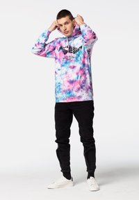 Felpa con cappuccio in un motivo tie-dye nei toni del rosa, blu e viola, con un design grafico nero sulla parte frontale. Indossata con jeans neri e sneakers bianche.