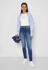 Blue and white vertical striped shirt, white cropped t-shirt with logo, dark blue skinny jeans, and white sneakers. Holding a dark blue cap.