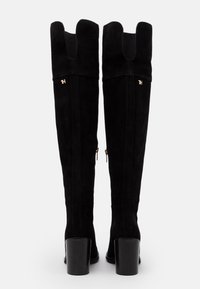 Botas de ante negras hasta la rodilla con un diseño de puño recortado único, que presenta acentos en dorado y un tacón grueso negro.