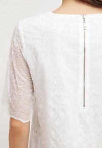 Valkoinen kirjailtu yläosa, jossa on läpikuultavaa kangasta monimutkaisilla kukkakuoseilla, pyöreä pääntie ja lyhyt vetoketjusuljenta takana.