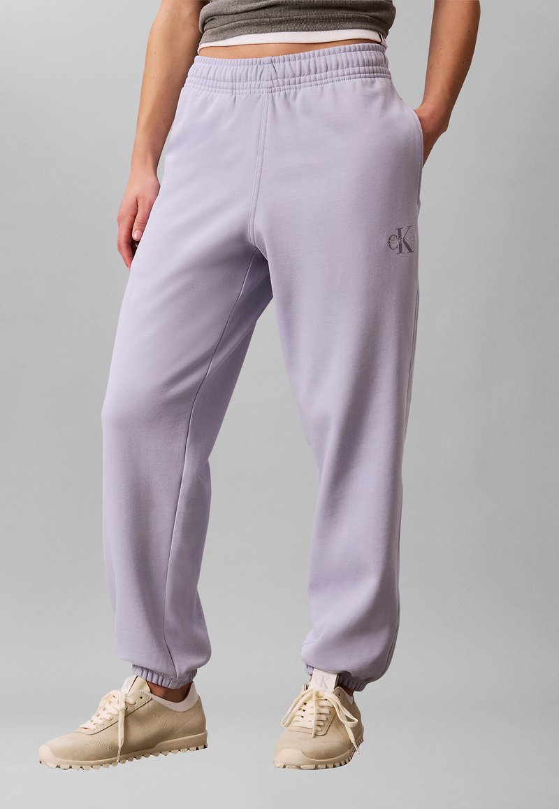 Personne portant un pantalon de survêtement Calvin Klein violet clair avec des poignets élastiques et des baskets beiges, debout devant un fond gris.