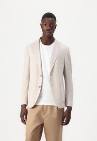 Béžový struktúrovaný blazer s klopami, dvomi gombíkmi a vreckami, nosený cez bielu košeľu a svetlohnedé nohavice.
