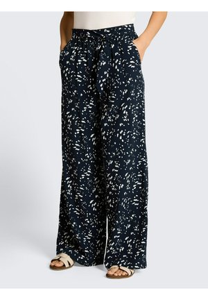 Marineblaue, weit geschnittene Hose mit weißen abstrakten Flecken, Taillengürtel zum Binden, getragen mit weißen Sandalen und Händen in den Taschen.